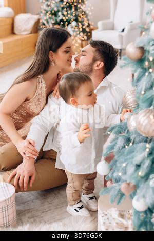Una famiglia felice trascorre del tempo di qualità accanto all'albero di Natale. I genitori condividono uno sguardo amorevole mentre il loro bambino esplora gli ornamenti e le decorazioni natalizie. Foto Stock