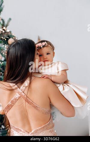 Un momento amorevole come madre condivide un bacio con sua figlia, entrambe vestite elegantemente, da un albero di Natale festoso adornato di ornamenti e luci. Foto Stock
