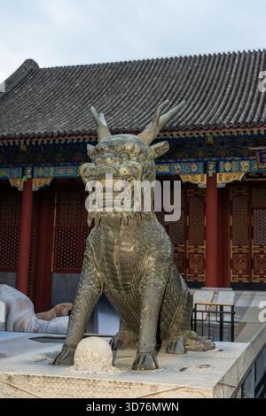 Una grande statua in bronzo di un Qilin (bestia mitica) sorge su un piedistallo in pietra davanti a un palazzo tradizionale cinese. La figura feroce protegge te Foto Stock