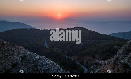 Veduta aerea di una strada tortuosa attraversa le colline dense e verdi della foresta mentre il sole tramonta sul lontano mare scintillante, Rodi, Grecia. Foto Stock