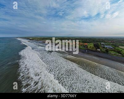 Vista aerea delle sabbie scure che incontrano le onde frizzanti a Seminyak, dove i resort si annidano tra la lussureggiante vegetazione di Seminyak, Bali, Indonesia. Foto Stock