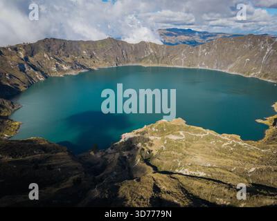 Vista aerea di uno splendido lago turchese annidato nel cratere di un vulcano dormiente, circondato da scogliere aspre e rocciose sotto un cielo striato di wispy c Foto Stock