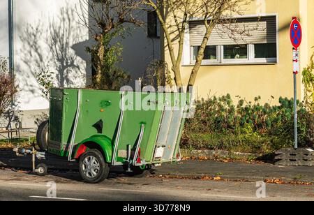 Accanto a un edificio residenziale si trova un rimorchio di colore verde brillante. Il rimorchio è posizionato sulla strada, circondato da alberi e erba. È sunn Foto Stock