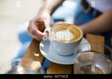 Una tazza di cappuccino con schiuma a forma di cuore tenuta per mano della donna su un tavolo di legno Foto Stock