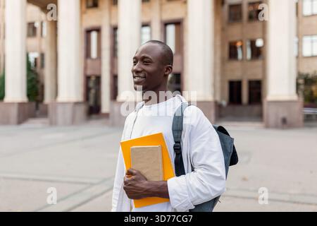 Gli studenti sorridono di fronte all'edificio universitario durante il giorno, tengono in mano libri e indossano uno zaino Foto Stock