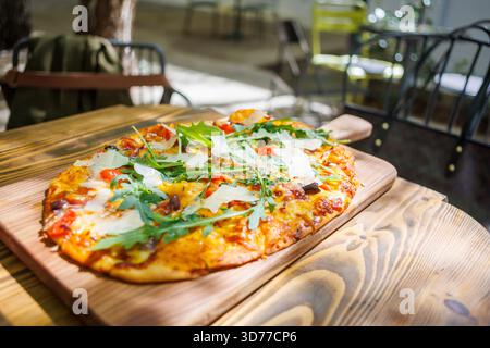 Una pizza appena sfornata condita con verdure e formaggio si siede su una tavola di legno in un accogliente caffè all'aperto in una giornata di sole Foto Stock