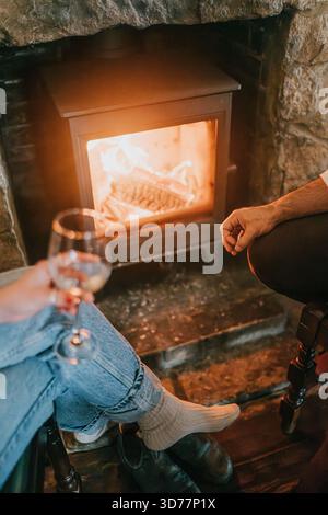 Un uomo e una donna sono seduti di fronte a un camino, tenendo dei bicchieri da vino. La scena è accogliente e intima, in quanto la coppia si diverte in compagnia l'un l'altro in un Foto Stock