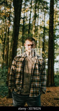 Un uomo è in piedi in una foresta, indossa una camicia a quadri e un jeans. Sta guardando in lontananza, forse perso nel pensiero. L'ambientazione della foresta crea Foto Stock