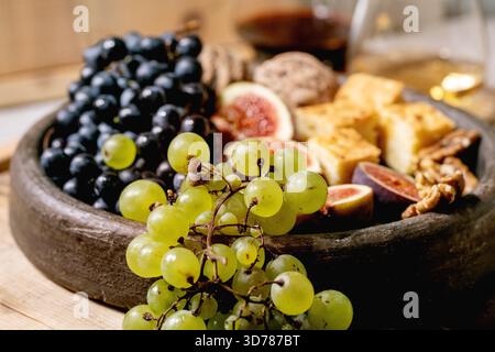 Antipasti con diverse uve, fichi, noci, pane, miele e formaggio di capra su un piatto di ceramica, serviti con bicchieri di vino rosso e bianco su un vecchio sfondo di legno. Primo piano Foto Stock