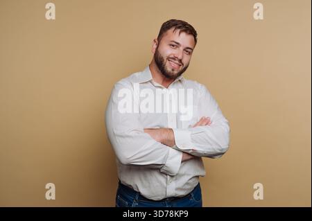 il ritratto di un giovane uomo sorridente posa con le braccia incrociate in una camicia bianca su sfondo beige Foto Stock