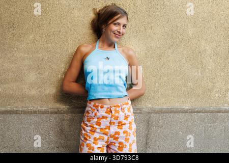 Giovane donna sorridente con sicurezza contro una parete testurizzata mentre indossa un top blu e pantaloni con motivi geometrici in un ambiente urbano Foto Stock