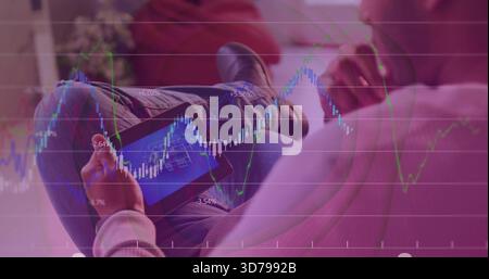 Tenendo in mano un tablet, un uomo che indossa una felpa leggera, un jeans che controlla le carte del mercato sovrapposte sul divano di casa Foto Stock