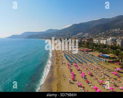 Alanya, Turchia - 13 giugno 2024: Ampia vista aerea di una trafficata spiaggia di sabbia fiancheggiata da centinaia di ombrelloni rosa e blu e lettini da sole, tramonti Foto Stock