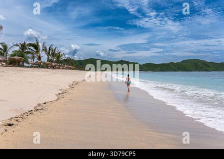 Lombok Indonesia – il turista cammina da solo sulla sabbia bianca vuota di una tranquilla spiaggia tropicale sotto un cielo azzurro. Foto Stock