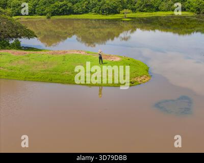 Vista aerea di una penisola verdeggiante dove una figura solitaria proietta un riflesso dorato sulle tranquille acque color caffè di El Encanto de Guanapalo, sa Foto Stock