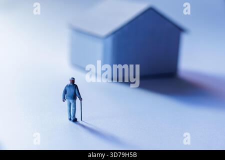 Figura di uomo anziano con canna si trova vicino a una piccola casa modello Foto Stock