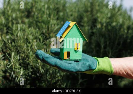 Piccola casa modello verde con pannelli solari tenuti in mano con guanti circondata dal verde in un giardino Foto Stock