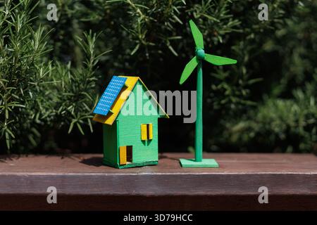Casa verde in miniatura con pannello solare e turbina eolica circondata dal verde in una giornata di sole Foto Stock
