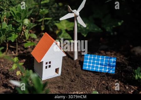 Casa in miniatura circondata da fonti di energia rinnovabili come turbine eoliche e pannelli solari in un giardino Foto Stock