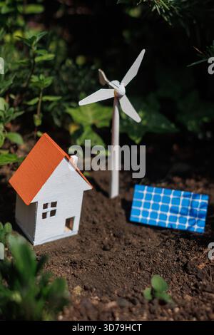 Casa in miniatura circondata da fonti di energia rinnovabili come turbine eoliche e pannelli solari in un giardino Foto Stock
