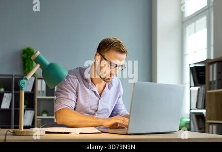 Revisore contabile mirato che esamina i documenti di controllo sul laptop in un moderno ambiente di lavoro Foto Stock