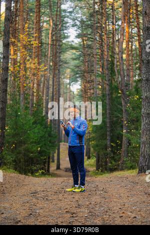 Uomo che indossa un abbigliamento sportivo e un cerchietto arancione in piedi su un sentiero nella pineta, che tiene e guarda il suo smartphone, magari controllando una mappa o un'app per il fitness Foto Stock