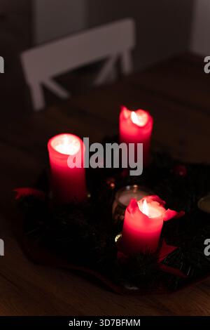 Una vista intima e poco illuminata di una corona dell'Avvento con tre candele a colonna rosse accese su una base verde scuro, che creano un'atmosfera calda e festosa Foto Stock