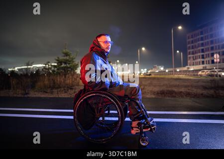 Un uomo con disabilità in sedia a rotelle si diverte a trascorrere del tempo all'aperto durante una passeggiata notturna in luce rgb al neon Foto Stock