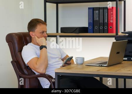 Un giovane uomo attento in un abbigliamento casual si siede pensiamente in un ufficio moderno, poggia il mento sulla mano, lo smartphone sull'impugnatura accanto al laptop aperto e alla scogliera fumante Foto Stock