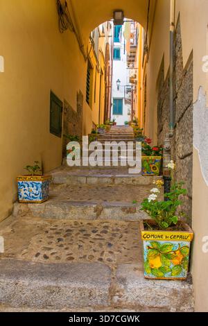 Una vista pittoresca di uno stretto vicolo a gradini a Cefalù, Sicilia, Italia. I gradini ripidi sono grandi blocchi di pietra usurati. Fioriere in ceramica maiolica Foto Stock