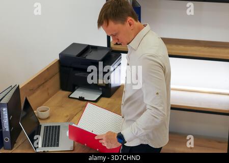 Giovane professionista in camicia leggera esamina documenti da legante rosso a una scrivania di legno con computer portatile e stampante nelle vicinanze. Foto Stock