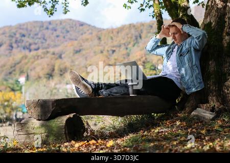 Giovane pensivo con le mani sulla testa, laptop e caffè accanto, contro gli alberi nella tranquilla foresta autunnale con montagne. Foto Stock