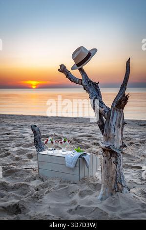 Picnic estivo sulla spiaggia con cappello sulla pelle, bicchieri da dessert e mare al tramonto. Concetto di relax per le vacanze. Foto Stock