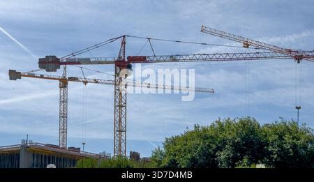 La gru domina il cantiere. I macchinari industriali in acciaio alto sollevano materiali pesanti con cavi e ganci, simboleggiando il progresso degli edifici urbani e. Foto Stock