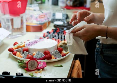 Mano della persona con carta di tessuto accanto a un piatto di dessert bianco circondato da frutta fresca assortita e frutti di bosco in un ambiente di cucina casalinga Foto Stock