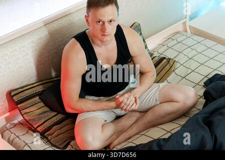 Un giovane uomo serio in abiti casual si siede sul letto con le mani strette, nel profondo del pensiero nell'ambiente domestico. Foto Stock