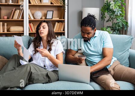 Coppia stressata che esamina le bollette della famiglia che effettuano il pagamento online a casa donna assegno documento mentre l'uomo inserisce i dettagli della carta di debito sul budget del notebook t Foto Stock