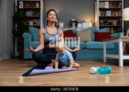Giovane madre che pratica yoga a casa con il bambino nelle vicinanze bilanciando recupero postpartum consapevolezza movimento delicato durante l'allenamento serale gioia legame con la famiglia Foto Stock