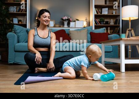 Giovane madre che pratica yoga a casa con il bambino nelle vicinanze bilanciando recupero postpartum consapevolezza movimento delicato durante l'allenamento serale gioia legame con la famiglia Foto Stock