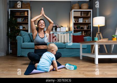 Giovane madre che pratica yoga a casa con il bambino nelle vicinanze bilanciando recupero postpartum consapevolezza movimento delicato durante l'allenamento serale gioia legame con la famiglia Foto Stock