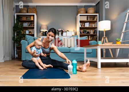 Giovane madre che pratica yoga a casa con il bambino nelle vicinanze bilanciando recupero postpartum consapevolezza movimento delicato durante l'allenamento serale gioia legame con la famiglia Foto Stock