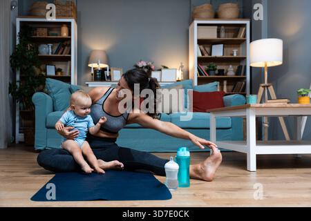 Giovane madre che pratica yoga a casa con il bambino nelle vicinanze bilanciando recupero postpartum consapevolezza movimento delicato durante l'allenamento serale gioia legame con la famiglia Foto Stock