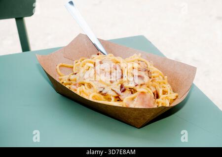 vista ravvicinata di cremosi spaghetti alla carbonara con pezzi di pancetta serviti in una ciotola usa e getta, su un tavolo verde all'aperto Foto Stock