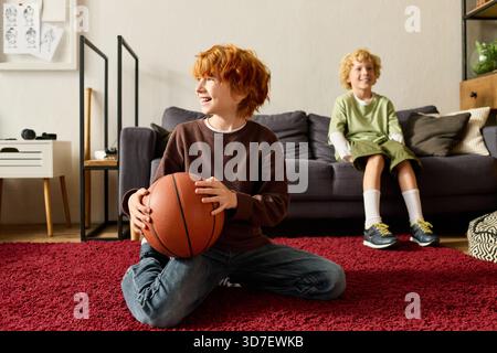 Due ragazzi allegri condividono un momento di divertimento a casa, uno che tiene una pallacanestro mentre è seduto sul tappeto. Foto Stock