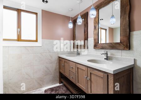 Un bagno elegante con doppio lavandino con specchio con cornice in legno naturale e luci a sospensione, accentuate da pareti piastrellate beige e una finestra in legno Foto Stock