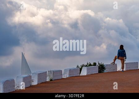 Una composizione drammatica con una figura solitaria in piedi su una barricata bianca, che si affaccia su una scena nuvolosa e suggestiva. Una strutturu bianca simile a una vela Foto Stock