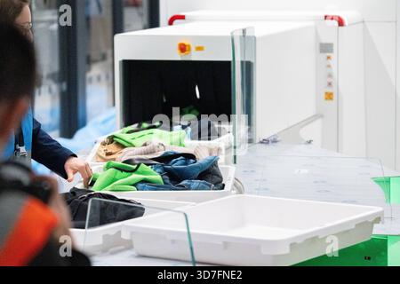 Baviera, Germania - 25 novembre 2025: L'abbigliamento e gli effetti personali di un viaggiatore aereo passano attraverso il controllo dei bagagli e di sicurezza. Il vassoio viene alimentato attraverso il moderno sistema a raggi X, mentre il personale di sicurezza controlla il contenuto per verificare la presenza di articoli proibiti. Il giorno delle operazioni di prova per il nuovo gate del Terminal 1 dell'aeroporto di Monaco. *** Kleidung und persönliche Gegenstände eines Flugreisenden passieren die Gepäck- und Sicherheitskontrolle. Die Wanne wird durch das moderne Röntgensystem geführt, während Sicherheitsmitarbeitende den Inhalt auf verbotene Gegenstände prüfen. AM Tag des Probebetriebs für den Foto Stock