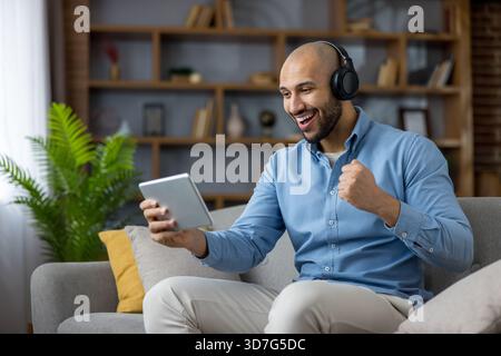 L'uomo calvo sul divano con le cuffie celebra la vittoria mentre guarda un tablet, stringendo il pugno di gioia durante i momenti rilassati di gioco domestico, streaming o videochiamate Foto Stock