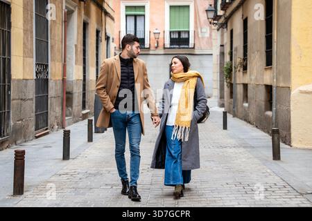 Una giovane coppia cammina mano nella mano lungo una stretta strada acciottolata a Madrid, vestita calorosamente con eleganti abiti invernali. Le loro risate riempiono l'aria Foto Stock