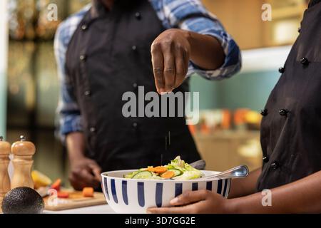 Giovane coppia afroamericana che prepara il cibo insieme nella loro cucina, aggiungendo un pizzico di sale a una fresca insalata estiva. Uomo e donna che godono di un'attività culinaria sana e organica. Foto Stock